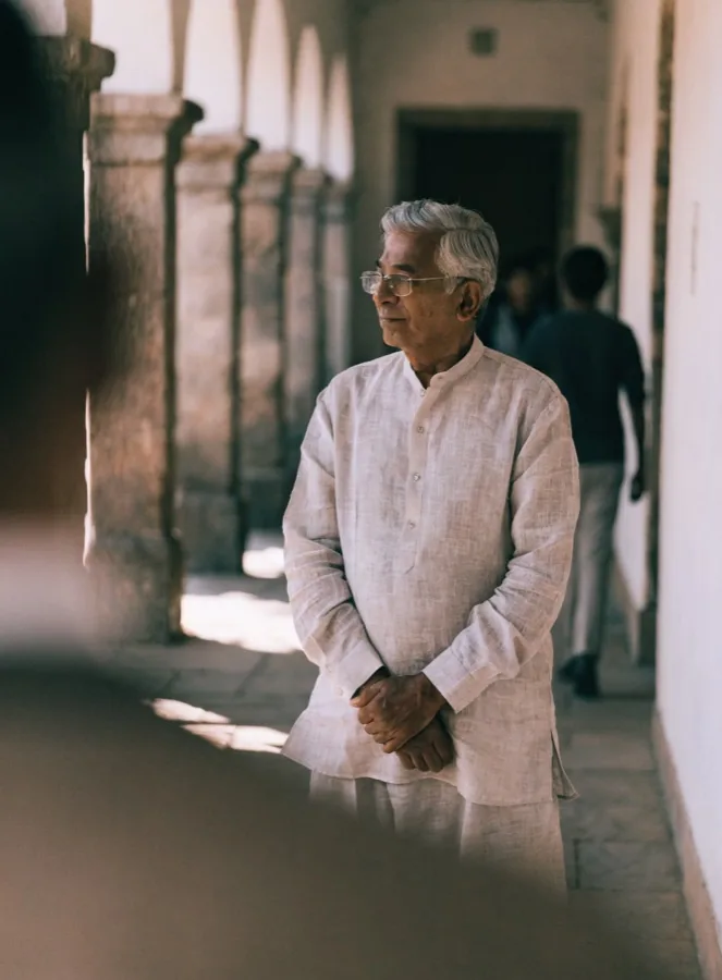 An older man in a white kurta standing in a sunlit colonnade